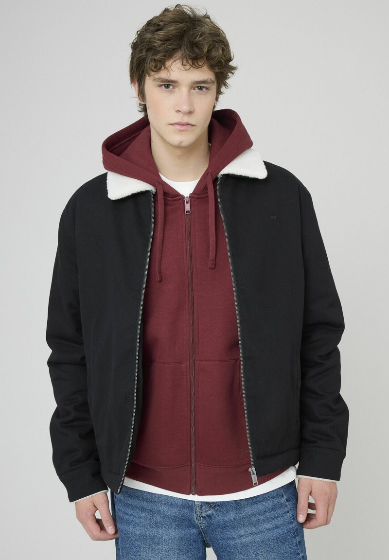 Veste aviateur noire avec col en fausse fourrure blanche sur un sweat à capuche zippé bordeaux. Le modèle porte un jean bleu. Texture lisse, design contemporain.