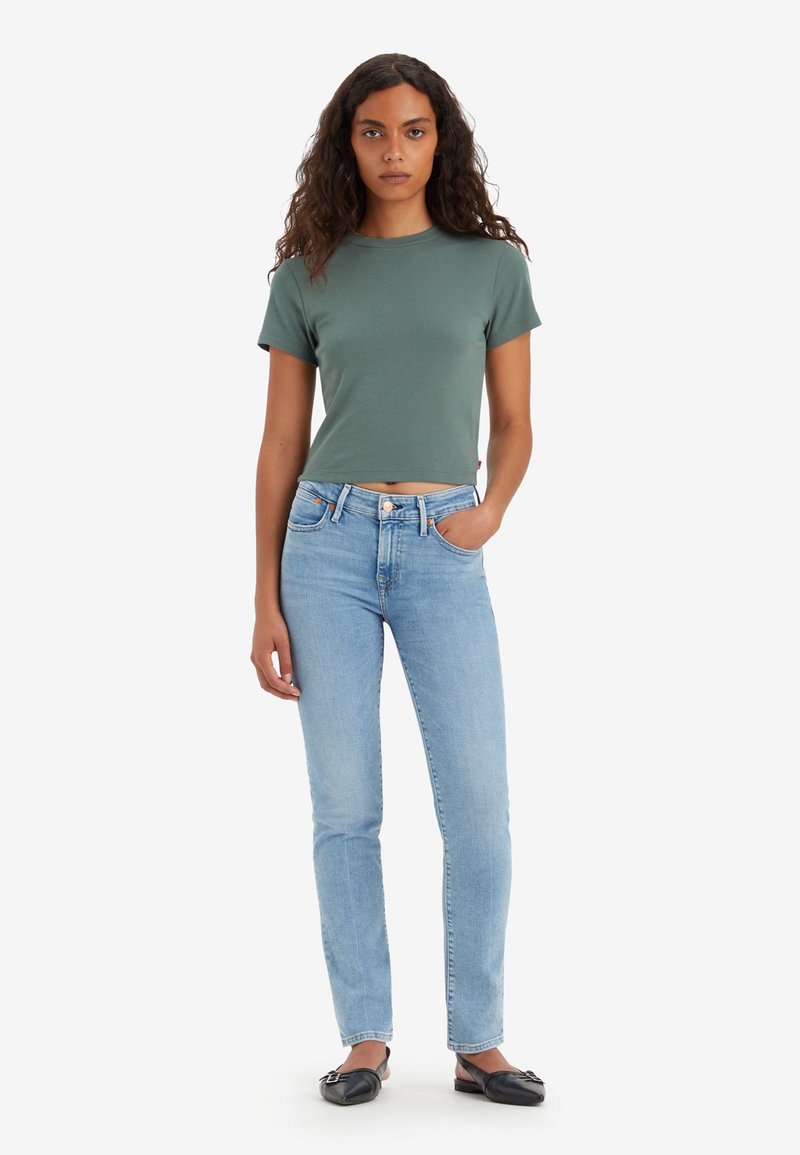 Camiseta verde recortada, diseño ajustado, combinada con jeans de pierna recta de color azul claro. El modelo lleva zapatos planos negros con detalle de lazo, sobre un fondo blanco liso.