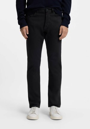 Mann trägt schwarze gerade geschnittene Hose, marineblauen Pullover und weiße Turnschuhe, stehend vor einem schlichten weißen Hintergrund.