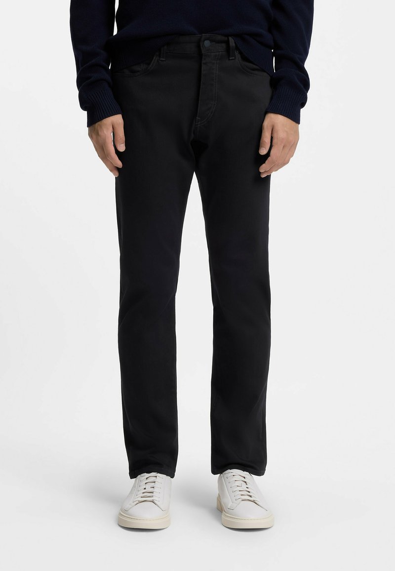 Mężczyzna ubrany w czarne spodnie o prostych nogawkach, granatowy sweter i białe trampki, stojący na tle jednolitego białego tła.