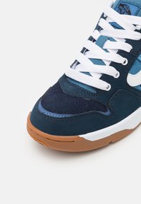 Sininen ja tummansininen mokkanahkakengät, joissa on valkoiset nauhat ja kuminen pohja. Ominaisuuksiin kuuluvat ompeluyksityiskohdat ja logo kielekkeessä.