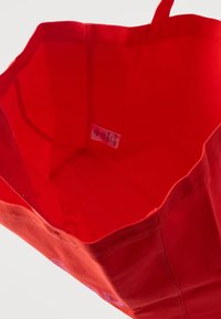 Κόκκινη υφασμάτινη τσάντα ώμου ανοιχτή στην κορυφή που δείχνει την επένδυση στο εσωτερικό και μια μικρή λευκή ετικέτα φροντίδας ραμμένη στην πλευρική ραφή.