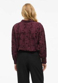 Blouse bordeaux avec broderies florales, manches longues et un design à dos froncé. Associée à un pantalon noir, offrant une coupe décontractée.