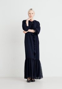 Vestido longo azul marinho com detalhe franzido na parte da frente, mangas longas e barra em camadas. Tecido leve com uma textura suave. Usado com sapatos pretos.