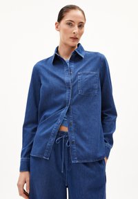 Camicia in denim blu scuro con bottoni, caratterizzata da un colletto, una tasca sul petto e maniche lunghe. Il tessuto ha una texture liscia.