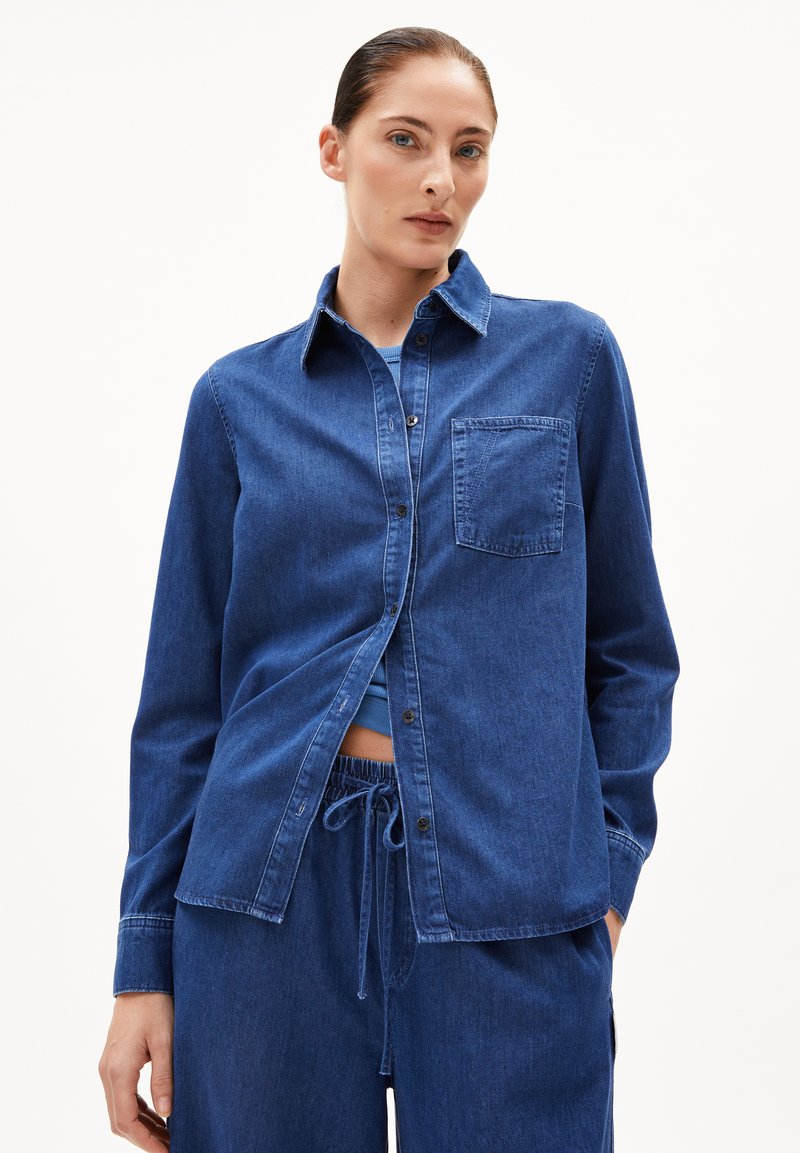 Camicia in denim blu scuro con bottoni, caratterizzata da un colletto, una tasca sul petto e maniche lunghe. Il tessuto ha una texture liscia.