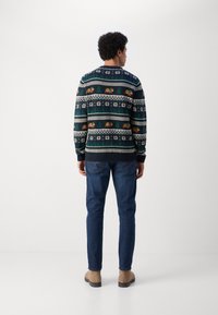 De gebreide trui heeft een meerkleurig patroon met dieren, sneeuwvlokken en geometrische ontwerpen op een marineblauwe achtergrond. Gecombineerd met blauwe jeans.