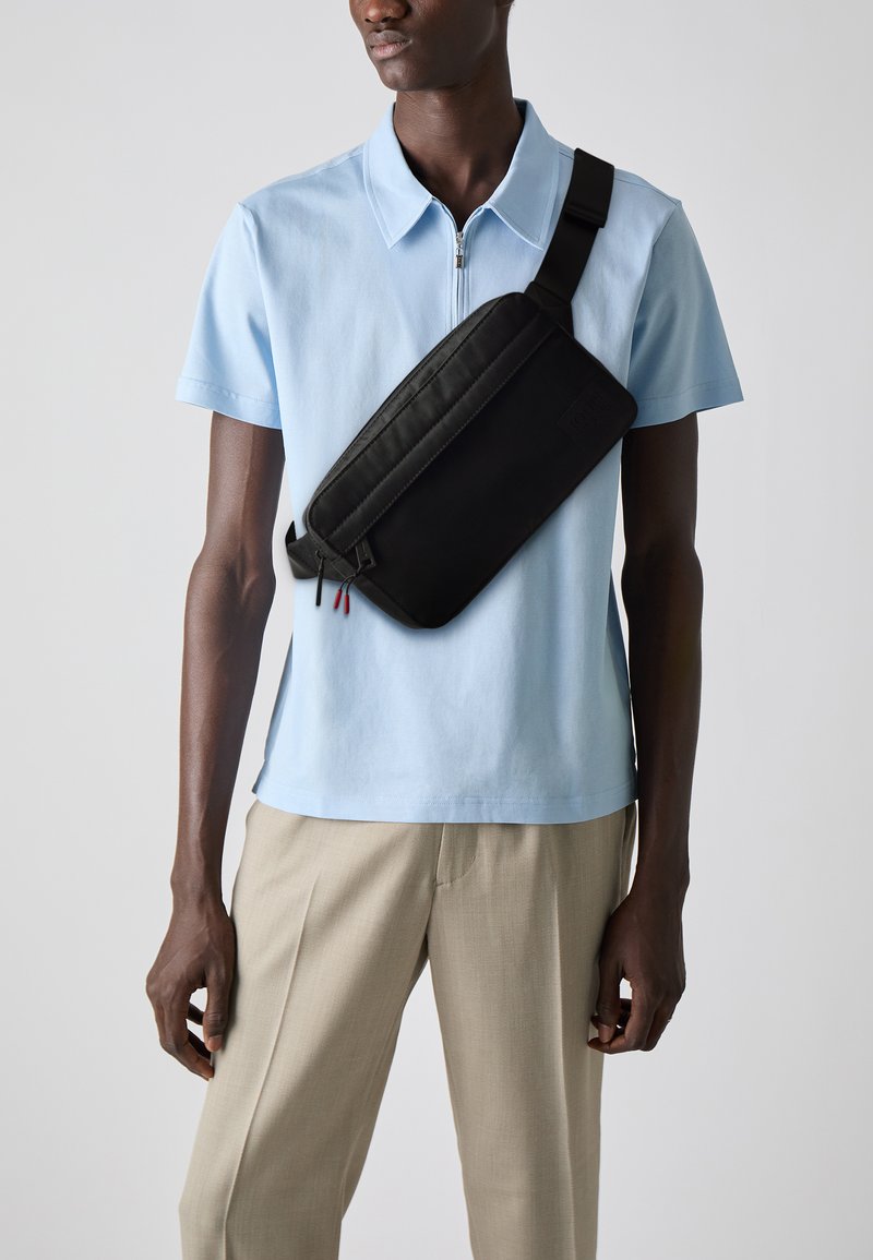 Uomo che indossa una camicia leggera azzurro chiaro a maniche corte con cerniera e pantaloni beige, con una borsa a tracolla nera attraversata sul petto.