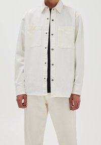 Jasbeżowa bawełniana kurtka koszulowa z dwoma kieszeniami na piersi, zatrzaskami i prostym krojem. Włożona na czarną bluzkę, w zestawie z jasnymi spodniami.