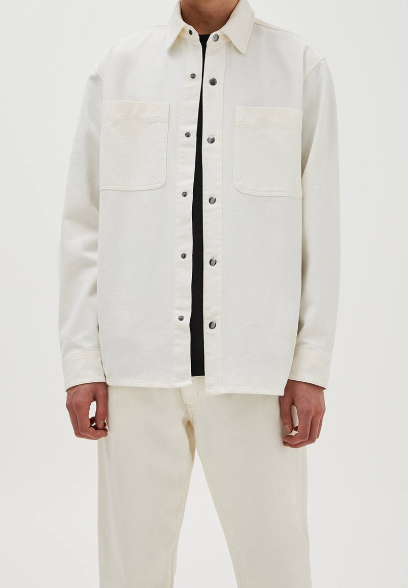 Jasbeżowa bawełniana kurtka koszulowa z dwoma kieszeniami na piersi, zatrzaskami i prostym krojem. Włożona na czarną bluzkę, w zestawie z jasnymi spodniami.