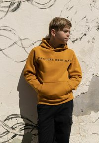 Mosterdgele hoodie met de tekst "DEE LUXE ORIGINAL" in het zwart, gecombineerd met zwarte cargobroek tegen een gestructureerde muur.