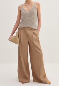 Top in maglia beige con scollo a V, abbinato a pantaloni a gamba larga marrone chiaro a vita alta. La modella tiene una borsa triangolare color tan.