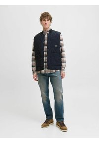 Quiltet navy vest med trykknapper og frontlommer båret over en ternet skjorte, parret med slidte blå jeans og brune snørebåndsstøvler.