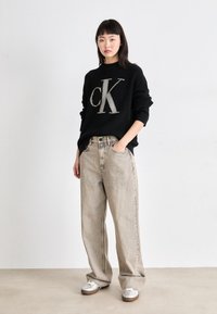 Črn pleten pulover z velikim kontrastnim logotipom "cK", v kombinaciji z svetlo sivo širokima denim hlačami in srebrnimi sproščenimi čevlji. Preprost dizajn.