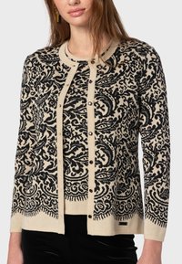 Beige kofta med intrikata svarta paisleymönster, rund hals, knäppning fram och ribbad nederkant. Mjuk stickad textur och figurnära ärmar.