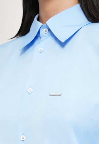 Light blue button-up shirt featuring a pointed collar, white buttons, and a small metallic logo on the chest pocket. Smooth fabric texture.