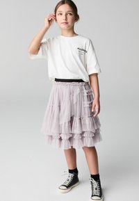 Vit t-shirt med svart logotyp, lager av tylkjol i ljus lila, elastiskt midjeband, svarta högskurna sneakers, bärs med svarta strumpor.