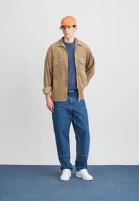 Camisa-jaqueta de corduroy castanha com dois bolsos no peito, t-shirt azul-marinho, jeans largos azuis, ténis brancos e um boné laranja. Em pé, contra um fundo simples.