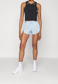 Schwarzes, kurzes Tanktop, hellblaue Shorts mit Kordelzug, weiße Sneaker und Socken. Nike-Logos auf dem Tanktop, den Shorts und den Socken.