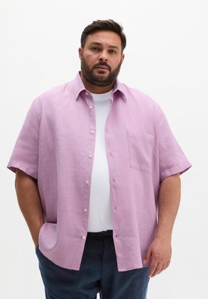 Mężczyzna z brodą, ubrany w jasnofioletową koszulę na krótkim rękawie zapinaną na guziki, na białej koszulce i ciemnych spodniach, stojący na jednolitym białym tle.