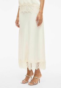 Kremhvit skjørtdel med blonderkant og intrikat blomsterdekor ved kanten. Går til under kneet, kombinert med sølvfargede sandaler med høye hæler og stropper.