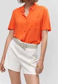 Krótki rękaw, jasnopomarańczowa koszula na guziki z kieszenią, w połączeniu z białymi szortami i jasnoszarym paskiem, na neutralnym tle.