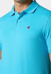 Tyrkysová polo košile z měkké, prodyšné látky; má límeček, krátké rukávy a malou červenou logo aplikaci na hrudi.