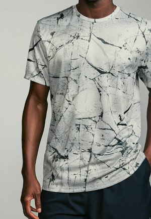 Mężczyzna ubrany w biały t-shirt z krótkim rękawem z czarnym abstrakcyjnym wzorem pęknięć i ciemne spodnie, z jedną ręką w kieszeni, a drugą w luźnej pozycji.