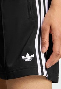 Pantaloncini atletici neri in tessuto liscio, con strisce laterali bianche e logo Adidas bianco vicino all'orlo. Tasca con cerniera visibile.