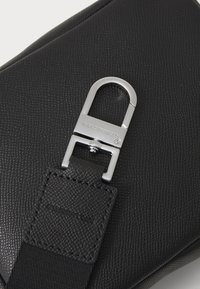 Sac en cuir noir texturé avec un clip et une sangle en métal argenté. Présente une forme rectangulaire et un logo de marque sur le matériel.