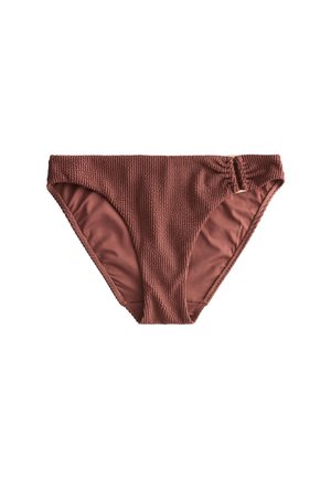 Kobieta w brązowym, teksturowanym bikini i bransoletkach spaceruje po słonecznej plaży z falami oceanu w tle.