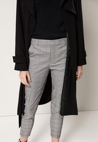 Zwarte lange jas met brede revers, gecombineerd met lichtgrijze geblokte broek, met een slim fit en cropped lengte, die diagonale lijnen accentueert.