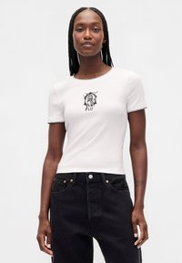 Hvit bomulls-T-skjorte med korte ermer, med et svart grafisk trykk og broderte detaljer rundt halsen, kombinert med svarte jeans.