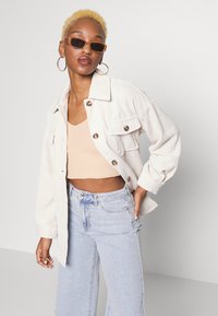 Casaco oversized de corduroy branco com botões, top cropped em bege claro e jeans de cintura alta em azul claro. Modelo a usar óculos de sol finos e brincos de argola.