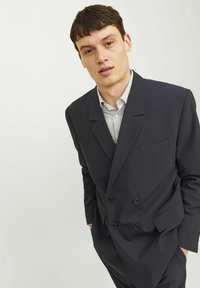 Marineblaue, doppelt geknöpfte Blazerjacke mit Revers, zwei aufgesetzten Taschen und schwarzen Knöpfen. Getragen über einem hellgestreiften Hemd. Glatte Textur.