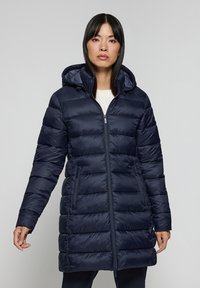 Marinblå puffjacka med huva, med dragkedja fram, horisontell sömnad och sidofickor. Slät, glänsande tyg med en något vadderad textur.