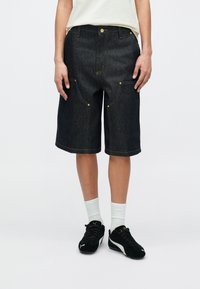 Oseba, oblečena v temne kavbojke kratke hlače do kolen, bele nogavice, črne superge znamke Puma in belo srajco, vtaknjeno noter, stoji na belem ozadju.