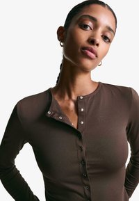 Bruine lange mouw top met een henley halslijn, voorzien van drukknoopjes en een getailleerd ontwerp. Het materiaal lijkt zacht en rekbaar te zijn.