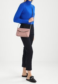 Žena v modrém roláku a černých kostkovaných zkrácených kalhotách, držící růžovou krosnu s řetízkovým popruhem a černými skládacími polobotkami.