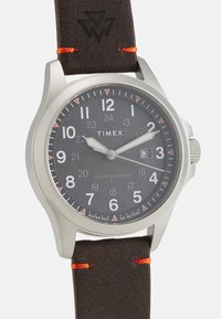 Montre à énergie solaire Timex avec un cadran rond noir texturé, boîtier en acier inoxydable argenté, chiffres blancs et un bracelet en cuir marron foncé avec des accents orange.