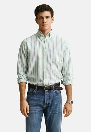 Jonge man die een lichtgroen gestreept overhemd met opgerolde mouwen draagt, een blauwe spijkerbroek, een gevlochten bruine riem en een zilveren polshorloge.