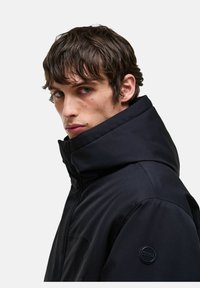 Fekete dzseki magas gallérral és texturált felülettel. Ujján foltlogó található, és modern, karcsú fazonban készült.