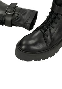Černé kožené kotníkové boty s kulatou špičkou, silnou podrážkou s hrubým vzorkem, předním šněrováním a nastavitelnou sponou na horní části holenní části.