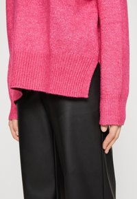 Kobieta w jasnoróżowym swetrze z dzianiny z bocznym rozcięciem i czarnych skórzanych spodniach, z ręką swobodnie opartą na boku na jasnym tle.