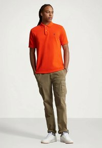 Homem em pé com as mãos nos bolsos de calças cargo caqui, vestindo uma camisola polo laranja brilhante e ténis brancos, olhando de lado.