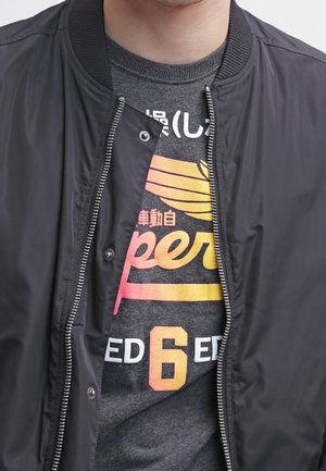 Schwarze Bomberjacke mit geripptem Kragen und Reißverschluss, getragen über einem dunkelgrauen T-Shirt mit buntem Grafiktext.