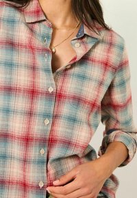 Red and blue plaid flannel shirt featuring a button-up front, rounded collar, and rolled-up sleeves. Light beige base with textured fabric.