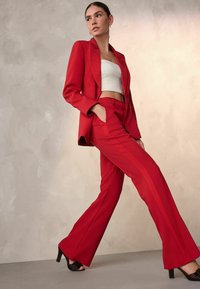 Punainen puku, jossa räätälöity, pitkähihainen bleiseri ja kapealahkeiset housut. Materiaali näyttää kuviolliselta, ja alla on sileä valkoinen crop-top.