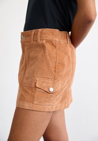 Personne portant un short en velours côtelé marron clair taille haute avec une poche à rabat latérale et un bouton argenté, associé à un haut noir.