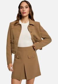 Chaqueta de mezcla de lana color marrón con botones dorados y grandes bolsillos, combinada con una falda marrón a juego que cuenta con un pliegue frontal y bolsillos.